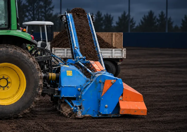 A tractor pulling a Fraze mower across a bare field, its conveyor lifting the stripped surface into a trailer