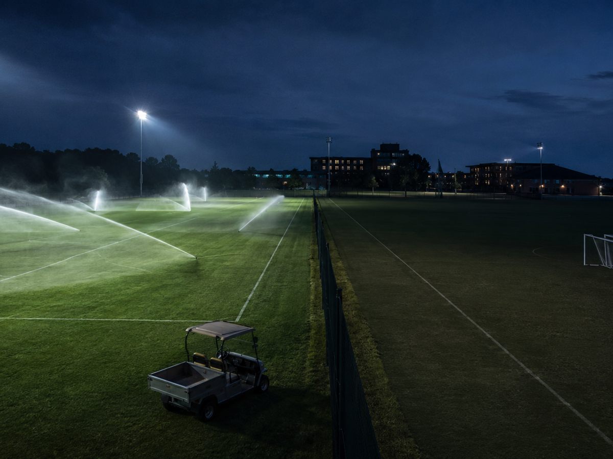 University practice fields at dusk with irrigation running and a utility cart parked at the fence line