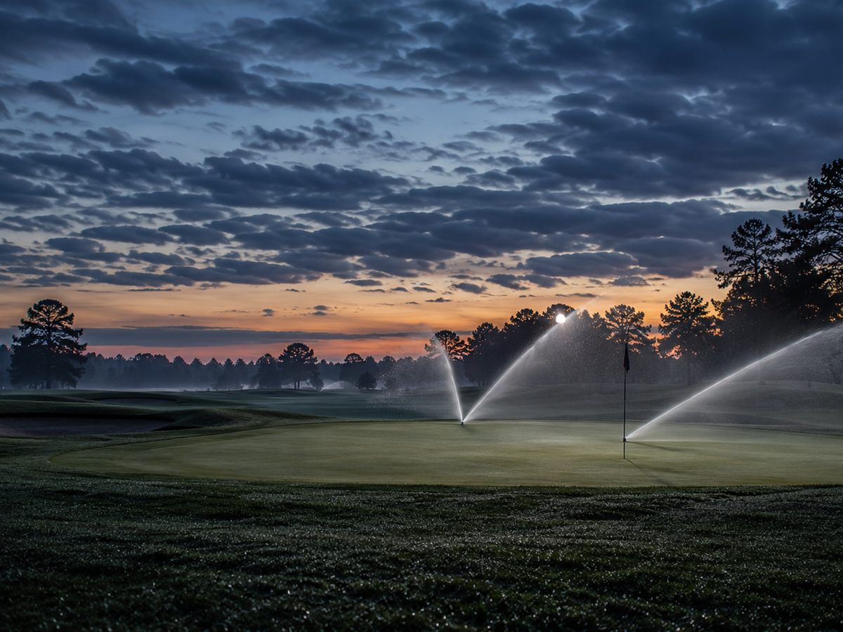 Golf green at dawn with sprinklers arcing near the flagstick in low mist